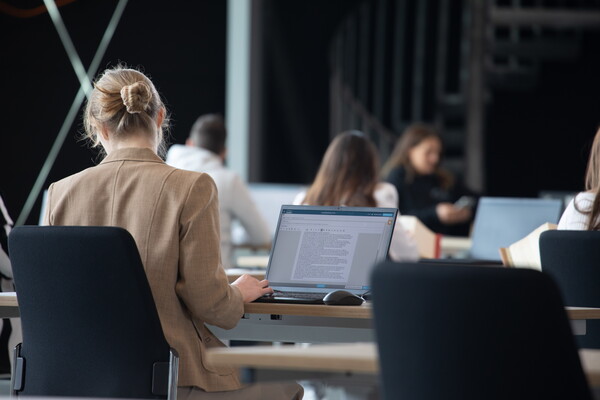 Eine Kandidatin im E-Exam sucht in Gesetzestexten nach Informationen, während sie eine Freitextaufgabe auf ihrem Laptop bearbeitet. Der Laptop zeigt die E-Exam-Aufgabe an.