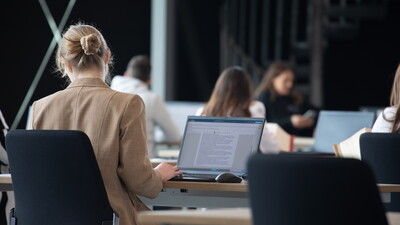 Eine Kandidatin im E-Exam sucht in Gesetzestexten nach Informationen, während sie eine Freitextaufgabe auf ihrem Laptop bearbeitet. Der Laptop zeigt die E-Exam-Aufgabe an.