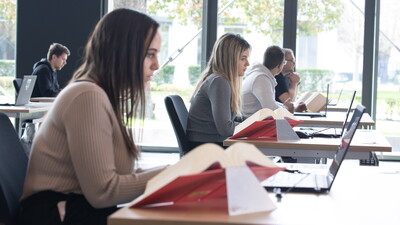 Seitlicher Blick auf Kandidat:innen , die während der E-Prüfung in einer Reihe sitzen, konzentriert arbeiten und sowohl Gesetzesbücher als auch Laptops vor sich haben.