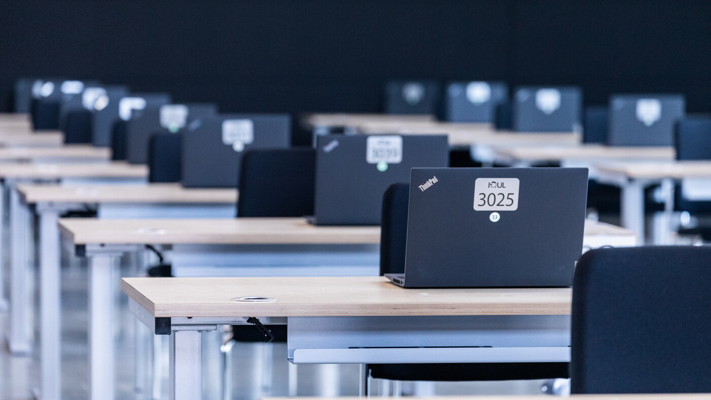 Ein ordentlicher Prüfungsraum mit sauber angeordneten Schreibtischen, auf denen aufgeklappte Laptops für die bevorstehende E-Prüfung bereitstehen.